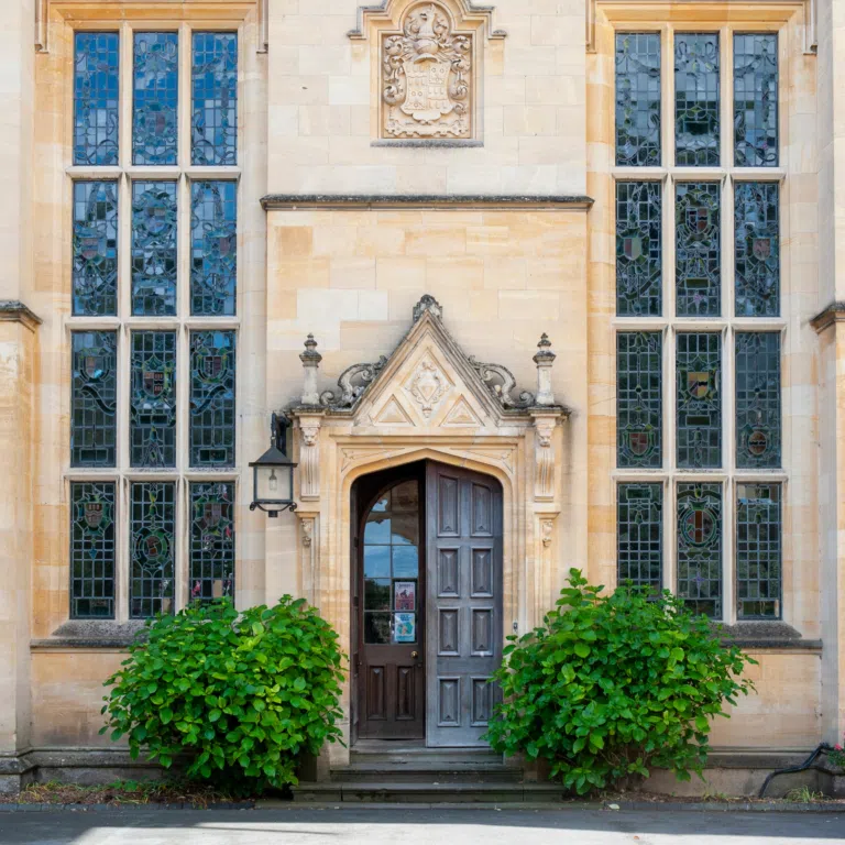 Beige stone building facade with tall stained-glass windows on both sides of a central arched wooden door and green shrubs in front.