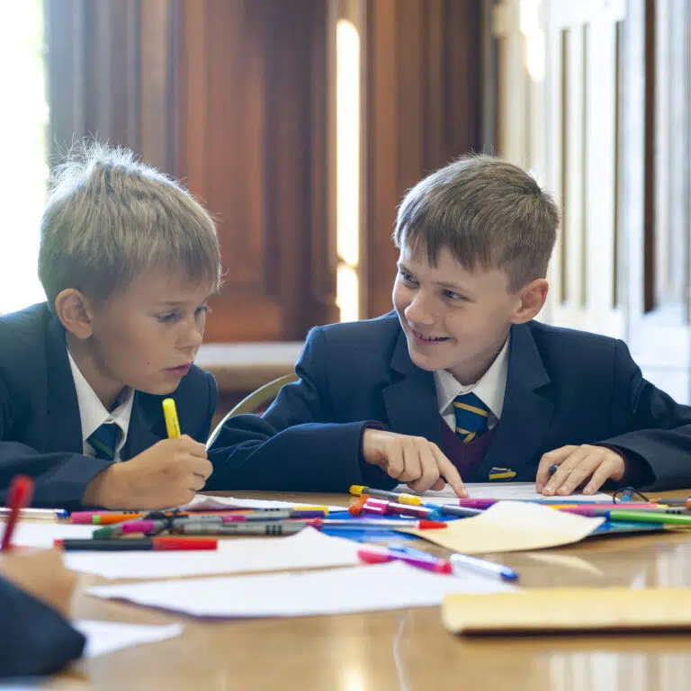 Two schoolboys in uniform drawing with markers at a table in a classroom. Bright colored pens scattered on the desk; warm wooden panel background.
