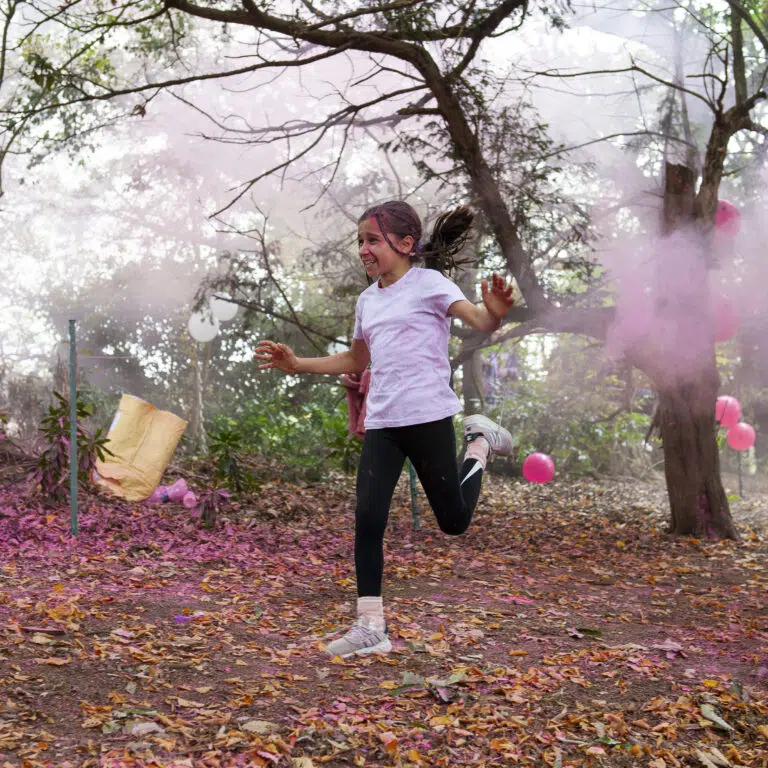 Girl in a white T-shirt runs and smiles as pink powder fills the air among autumn leaves in a park event.
