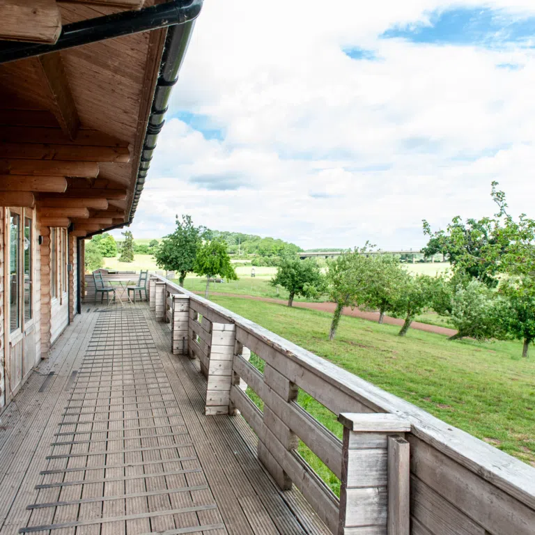 Wooden balcony of a rustic cabin with railing, overlooking green fields and trees under a cloudy blue sky.