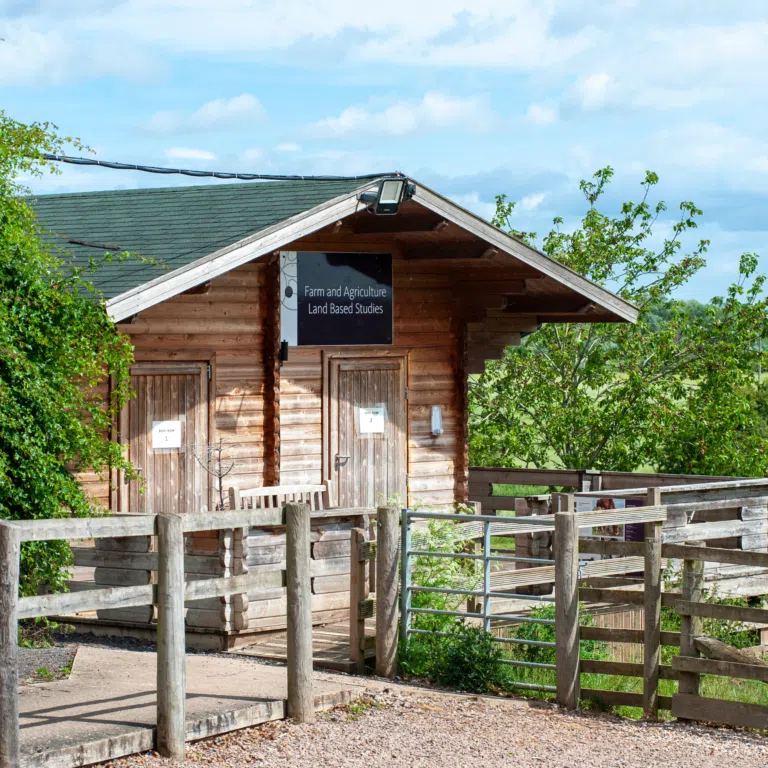 Wooden cabin with a sign reading 'Farm and Agriculture Land Based Studies' under a blue sky.