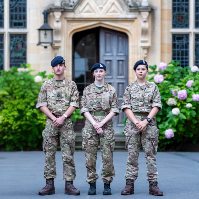Three soldiers in camouflage uniforms stand side by side outside a stone building with greenery and pink flowers in the foreground.