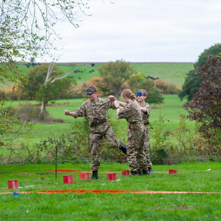 Soldiers in camouflage uniforms participate in a blindfolded teamwork exercise in a grassy field with red plastic cups and orange tape markers on the ground.