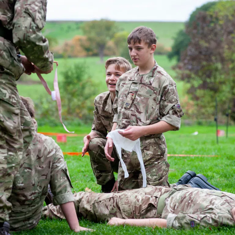 Two soldiers in camouflage assist a teammate lying on the grass during a field first-aid drill.