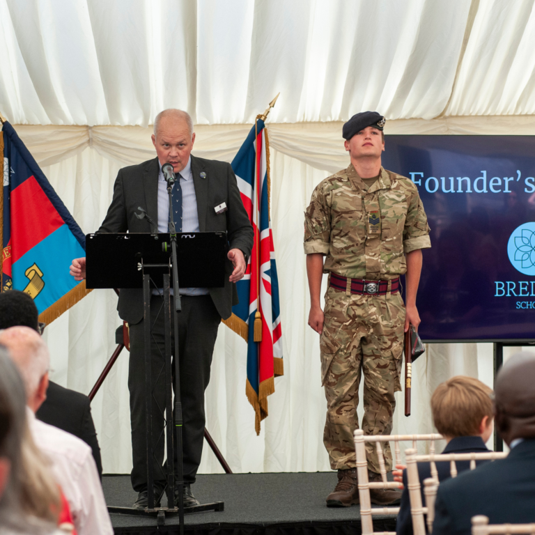 Man in a suit speaks at a lectern with a microphone, while a cadet in camouflage stands beside flags at a formal ceremony.