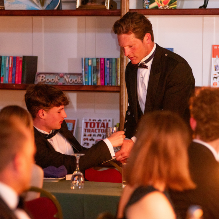 A man in a tuxedo leaning over a table and handing something to a seated person in a crowded bookstore or cafe setting.