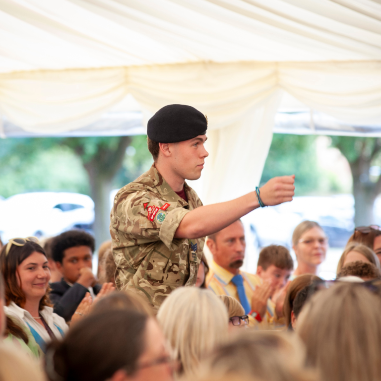 Young man in camouflage uniform with a black beret standing in a crowded outdoor audience under a tent.
