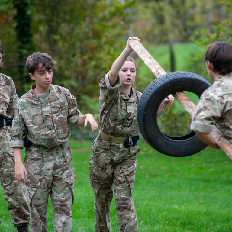 Cadets in camouflage uniforms outdoors, holding a wooden log and a large tire as part of a teamwork activity.