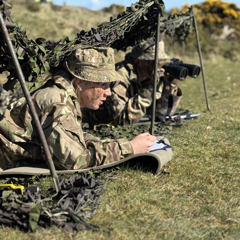 Soldier in camouflage gear sits on the ground writing in a notebook during a field training exercise under camouflage netting.
