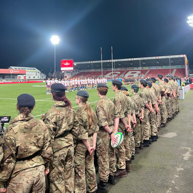 Line of soldiers in camouflage uniforms and boots standing on a rugby field, each holding a rugby ball, during a ceremony at a stadium.