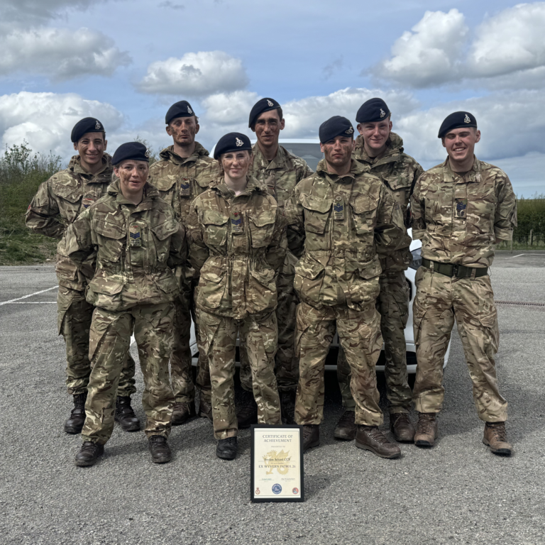 Seven soldiers in camouflage uniforms and black berets pose outdoors with a framed certificate on the ground in front.