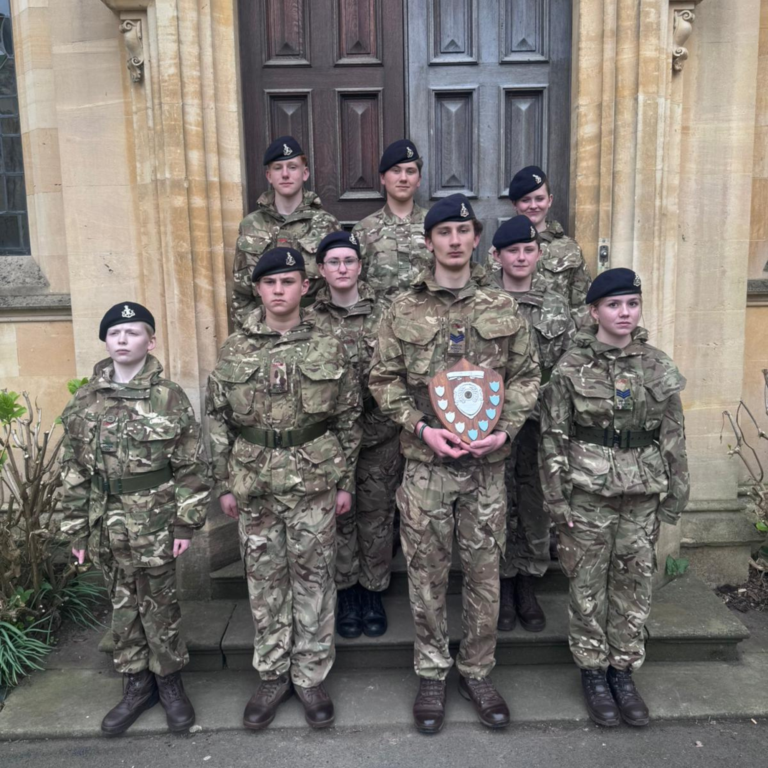 Group of cadets in camouflage uniforms and berets posing in front of a stone building doorway; one holds a shield with crest visible.