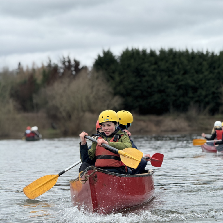 Child in a red canoe wearing a yellow helmet paddles on a lake, splashing water; other canoeists and trees in the background.