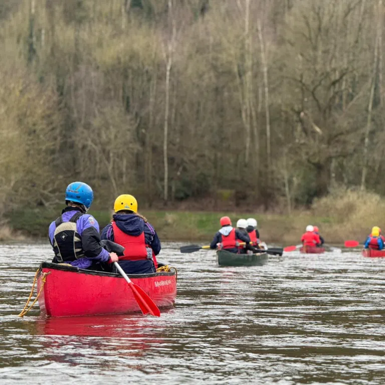 Group of people in canoes wearing helmets and life jackets on a river, paddling together with a forested bank in the background.