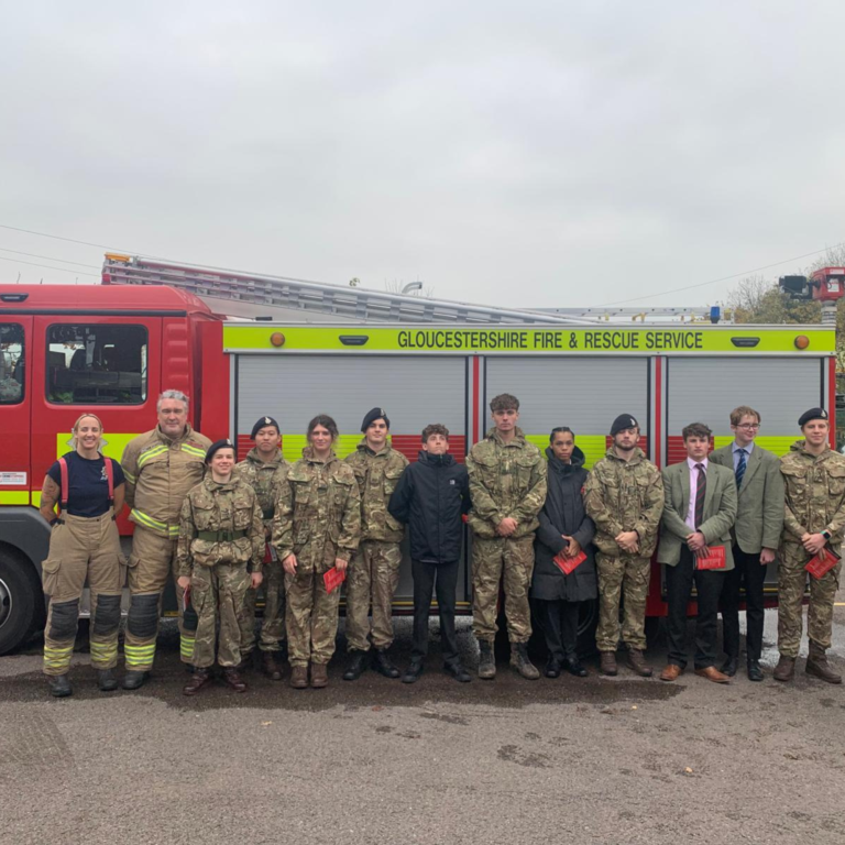 Group of firefighters and staff standing in a line in front of a red Gloucestershire Fire & Rescue Service fire engine, outdoors under a cloudy sky.
