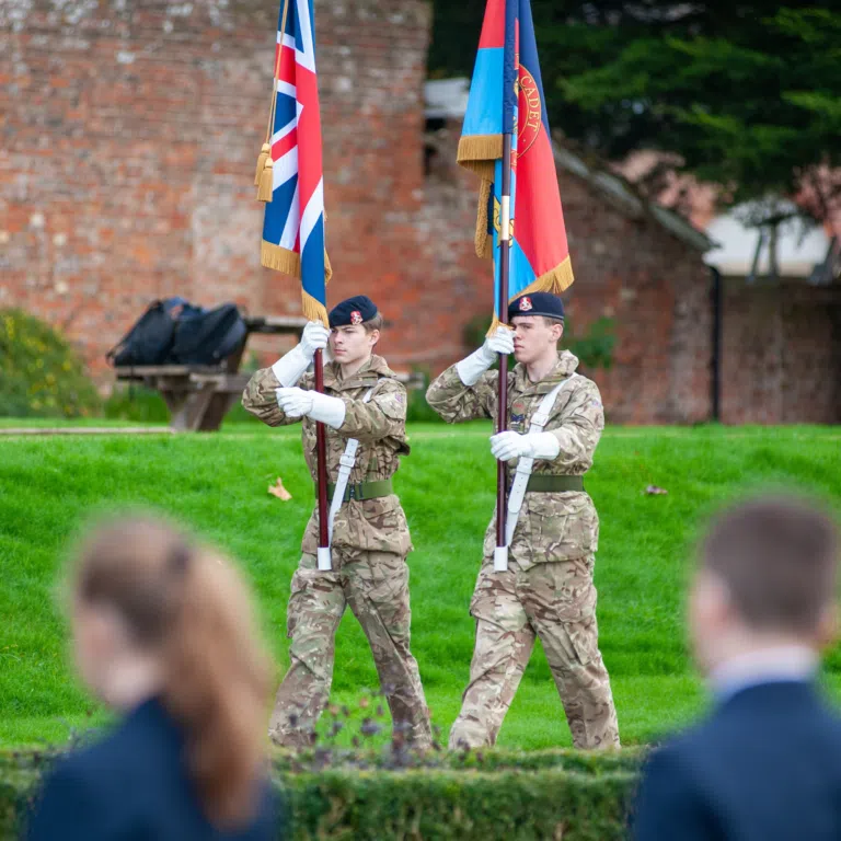 Two soldiers in camouflage uniforms hold ceremonial flags during an outdoor color guard event on a grassy area.
