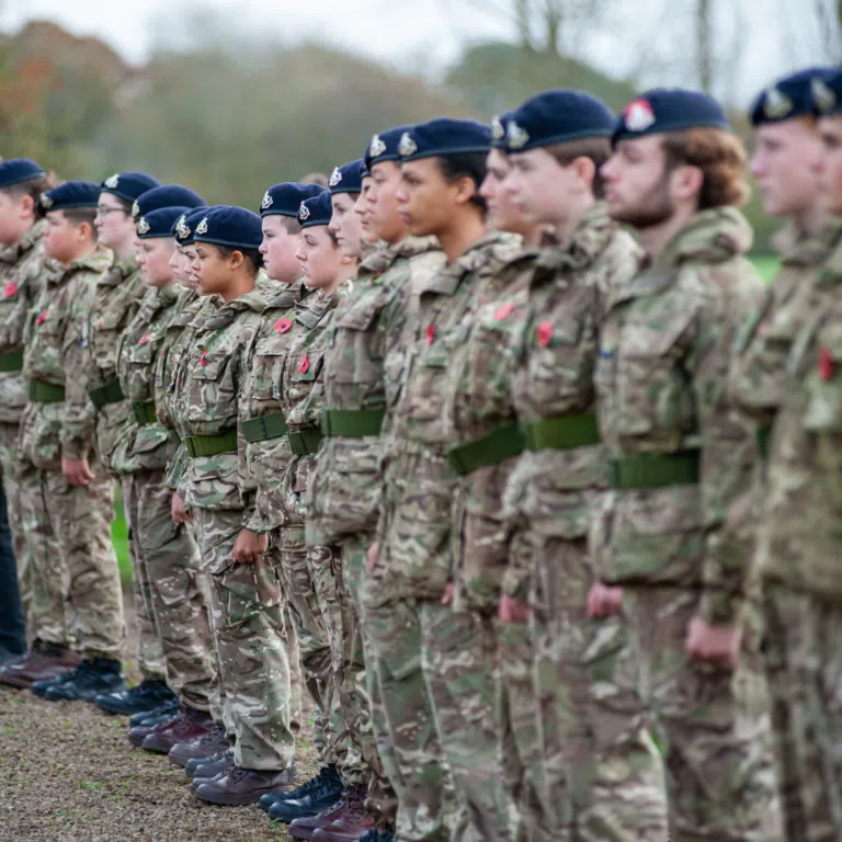 Line of soldiers in camouflage uniforms standing at attention outdoors, wearing navy berets with insignia.