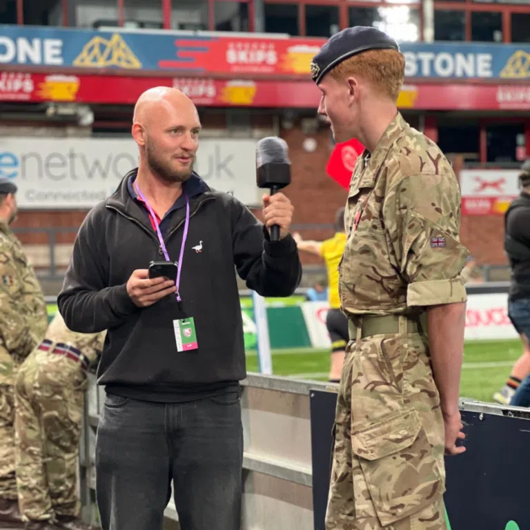 Interviewer in black hoodie holding a microphone speaks to a uniformed serviceman in camouflage at an event.