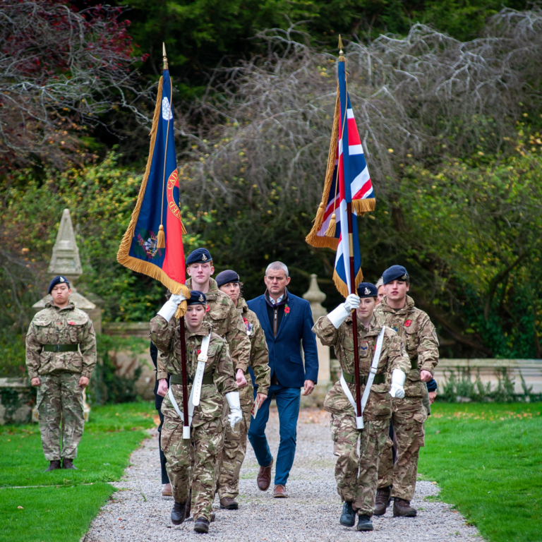 Soldiers in camouflage march along a gravel path, carrying ceremonial flags with gold fringe and tassels.
