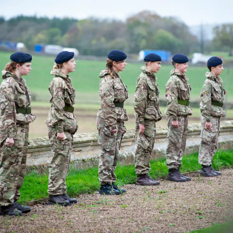 Line of soldiers in camouflage uniforms standing in a field, facing left, wearing blue berets.