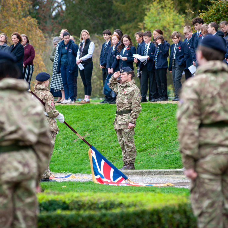 Outdoor ceremony with students in blue blazers and ties standing in a line as a soldier in camouflage salutes in the foreground, a Union Jack flag on the ground nearby.