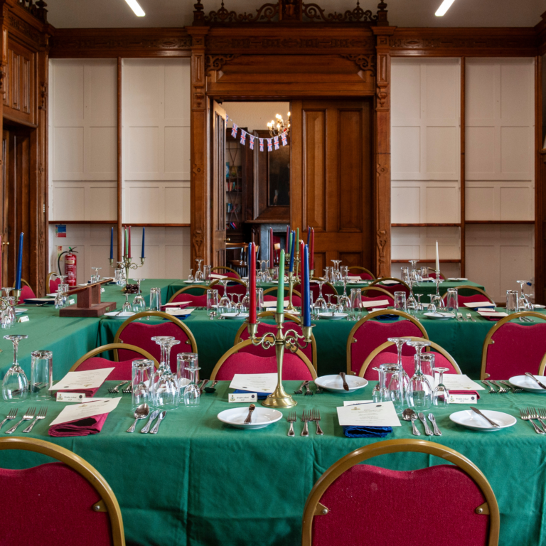 Formal dining room with green tablecloths, red chairs, gold candelabras, and Union Jack bunting in the doorway.