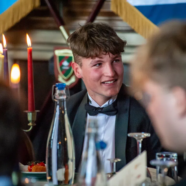 Young man in a tuxedo smiling at a candlelit celebration, with a cake and decorations in the foreground.