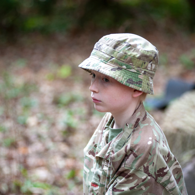 Young person wearing camouflage military uniform and hat, outdoors in a forested area, looking to the left to the side of the frame.