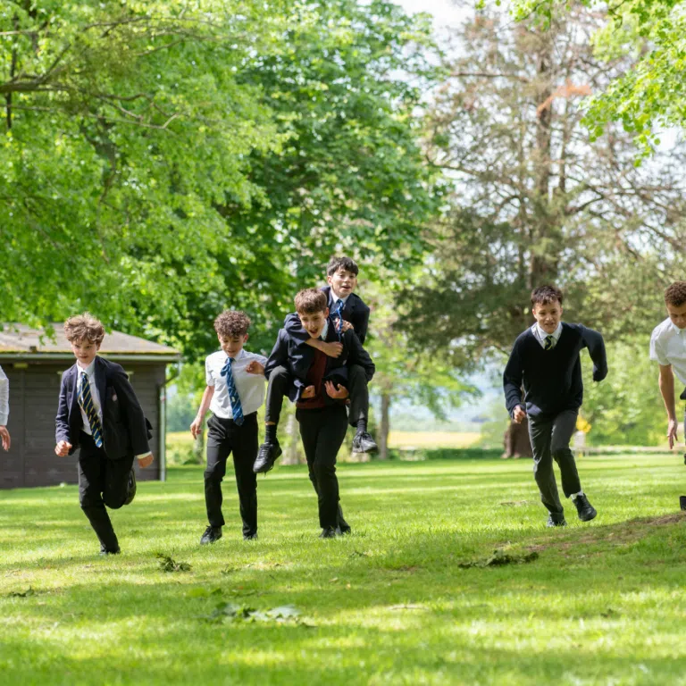 Group of schoolboys in uniforms sprinting across a sunny green park, with one boy being carried on another's shoulders while others run ahead and behind them.