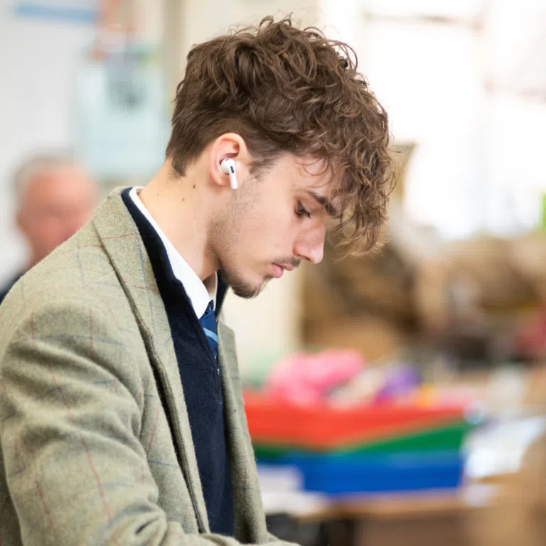 Person with curly hair wearing white wireless earbuds, looking down in a bright indoor setting (likely a classroom or cafe).