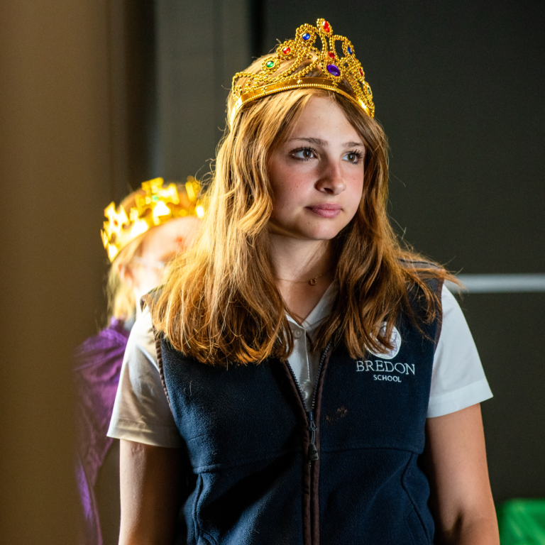 Girl wearing a gold crown with colorful gems, looking slightly to the side, in a navy vest over a light shirt (school uniform).