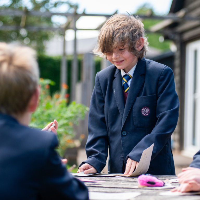 Schoolboy in a navy blazer and striped tie leans over a table outdoors, handling papers while classmates blur in the background.