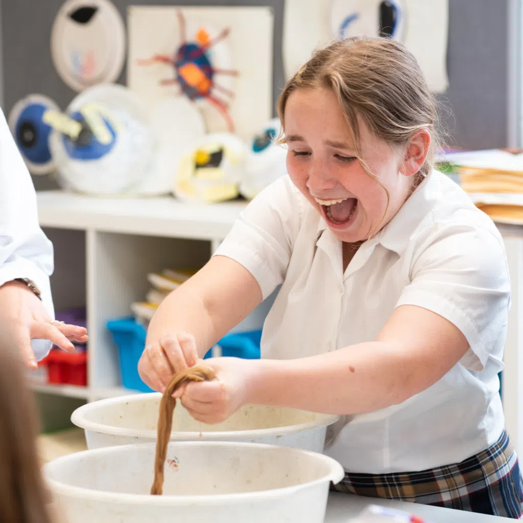 Person in a white shirt leaning over a large white bucket, kneading or pulling a dough-like substance with both hands in a classroom or workshop setting.