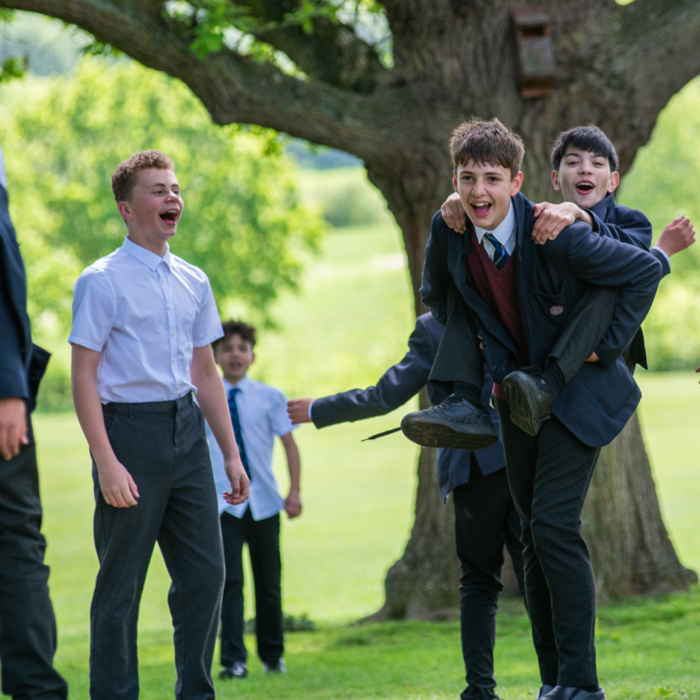 Schoolboys in uniform laughing outdoors as one student is carried on teammates' shoulders near a tree in a sunny park.