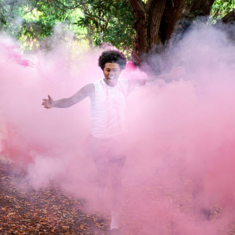 Person running through a forest as pink powder fills the air around them, smiling.