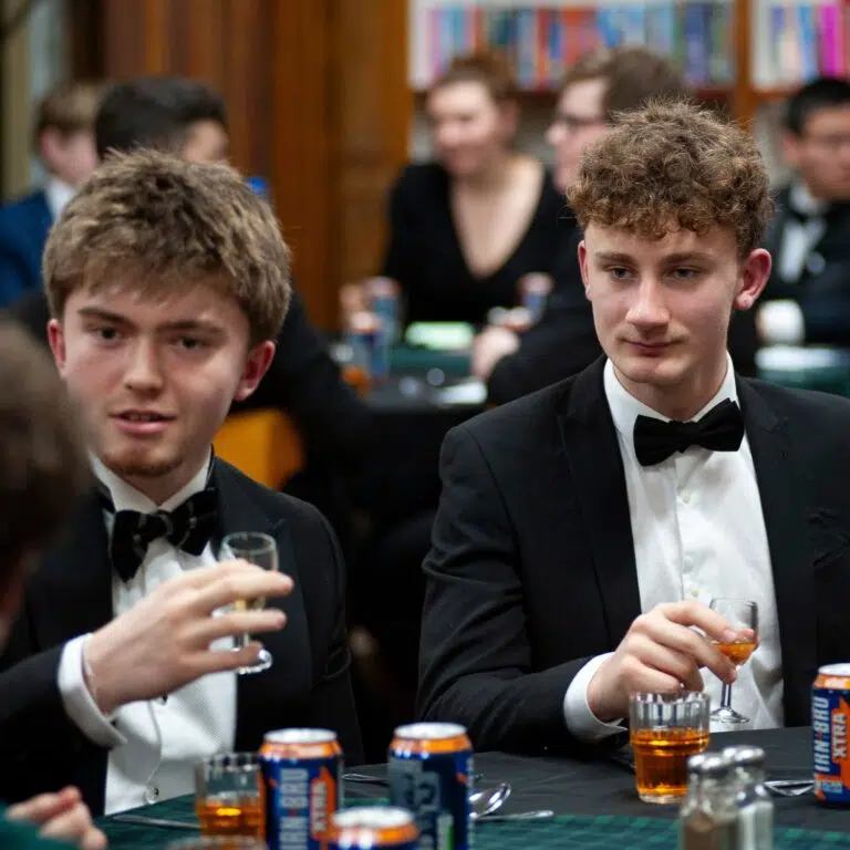 Two men in tuxedos sit at a table with drinks, foregrounded, while others chat in a blurred background at a formal event.