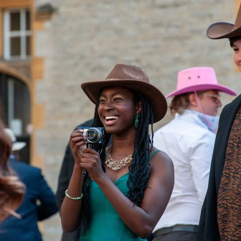 Smiling woman in a teal dress wearing a brown hat, holding a small camera outdoors at a festive gathering.