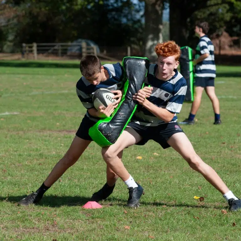 Two youth rugby players in blue-and-white striped jerseys struggle for the ball on a grassy field, with a bright green tackle shield nearby and a pink cone on the ground behind them.