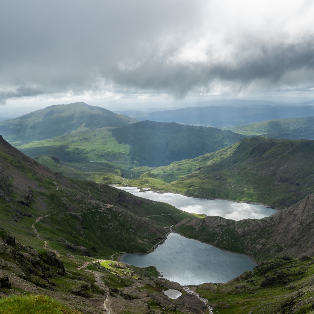 Mountain valley with a winding road along a blue lake, green hills, under a heavy gray clouded sky.