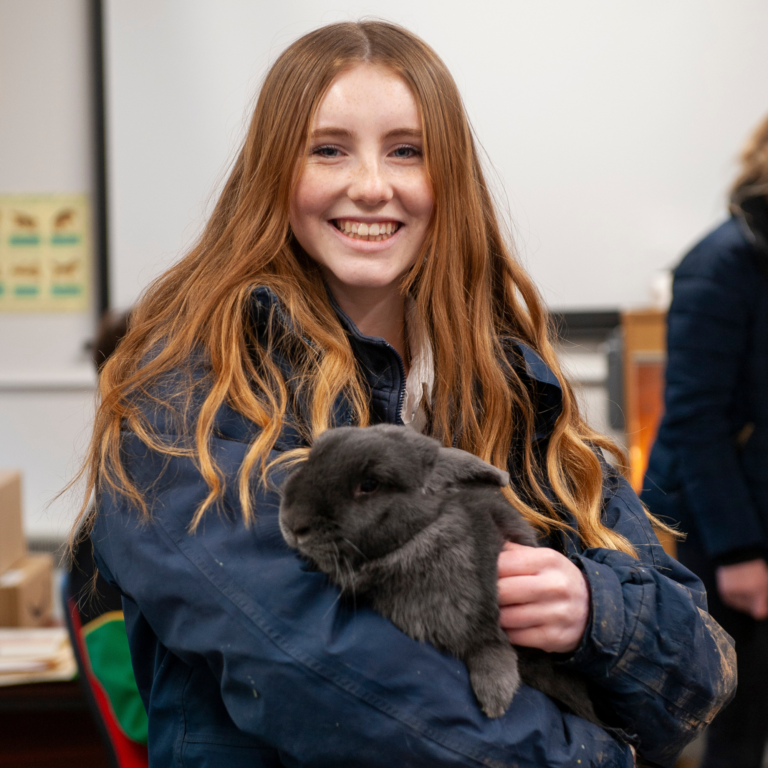 Smiling girl with long red hair holding a black puppy in a blue jacket indoors.