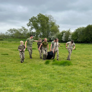 Group of people in camouflage clothing in a grassy field under a cloudy sky, with a folded parachute bag on the ground in front of them.