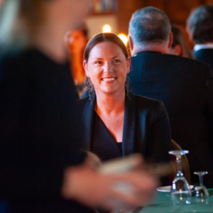 Smiling woman in a black outfit at a dimly lit social gathering, with blurred guests in the background and a glass on a green table in the foreground.
