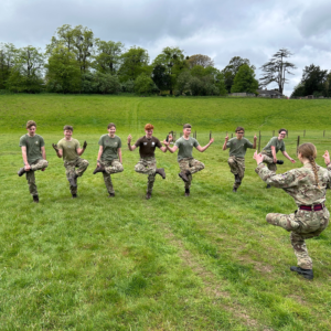 Group of cadets in camouflage uniforms jumping in a grassy field with tree-dotted hill and cloudy sky in the background.