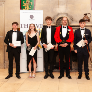 Group of five well-dressed students posing with certificates at a school awards event indoors, with a banner in the background.