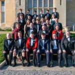 Group of students and adults in formal attire posing on stone steps outside a beige building for a class photo.