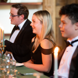 Woman in a black dress smiles at a candlelit dinner, flanked by two suited men at the table.