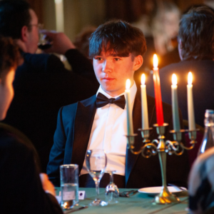 Young man in a tuxedo and bow tie at a candlelit formal event, with candles glowing on the table in the foreground.