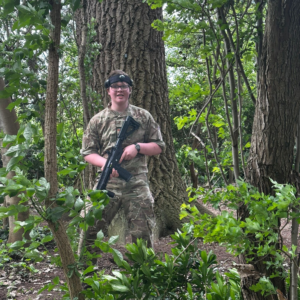Person in camouflage uniform stands among trees, holding a rifle pointed downward.
