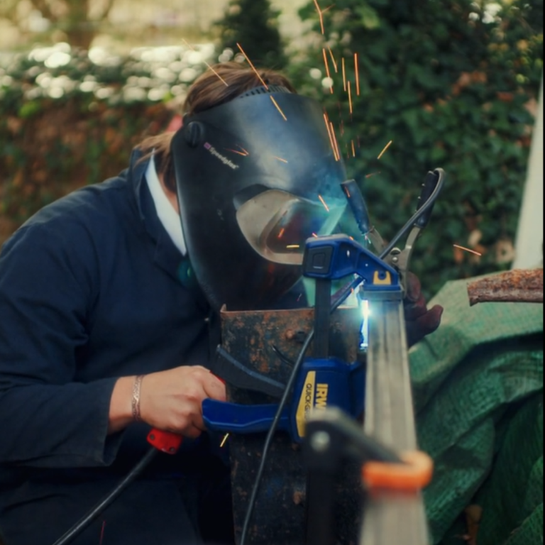 Welder wearing a protective helmet and blue jacket, sparks flying as they weld metal outdoors