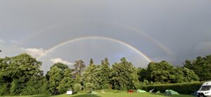 Double rainbow arches over a forested campsite with tents and a white van on a grassy field.
